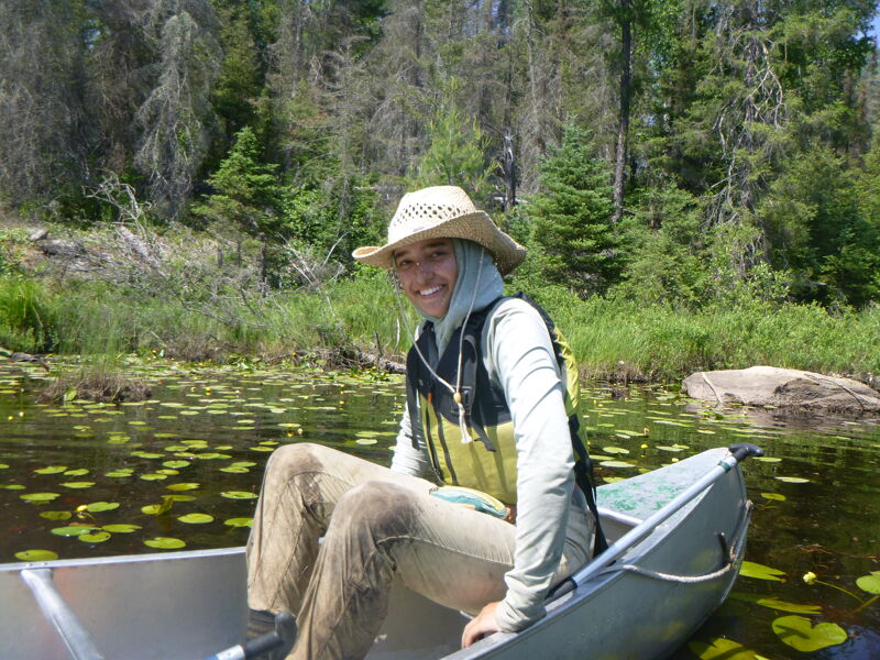 A person wearing a straw hat, a light-colored long-sleeved shirt, and a life vest is sitting in a canoe on a body of water covered with lily pads. The person is smiling and appears to be enjoying a peaceful day on the water. The background shows a lush green forest with tall trees, suggesting a natural and serene environment. The canoe is silver, and the water reflects the surrounding greenery.